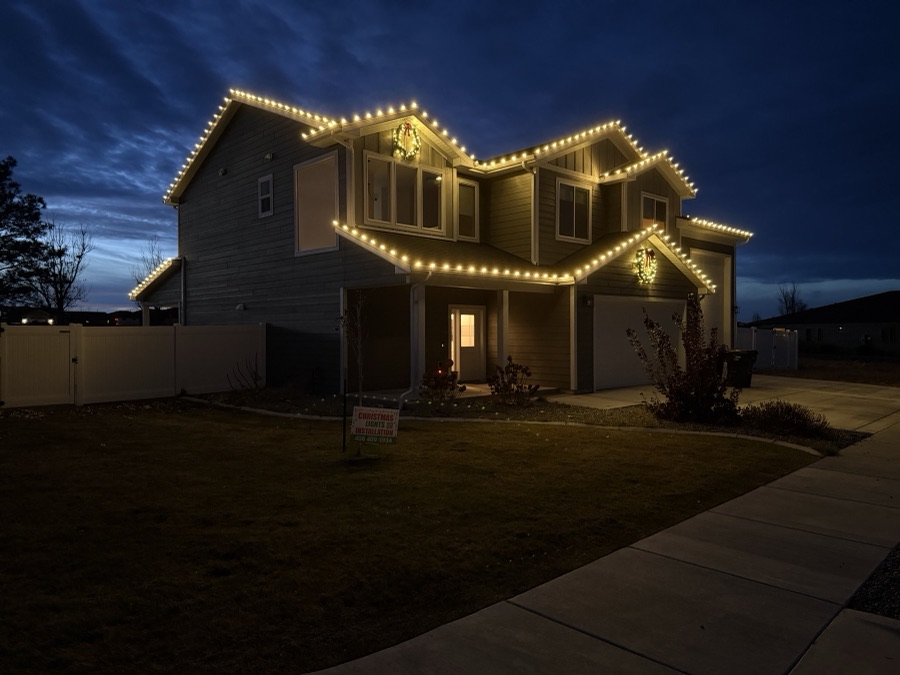 Two-story Montana home outlined in warm white roofline lights with lit wreaths at dusk
