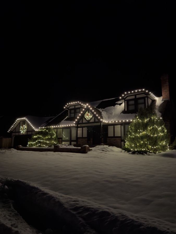 Tudor style home in deep snow with warm white roofline lights and two lit pine trees