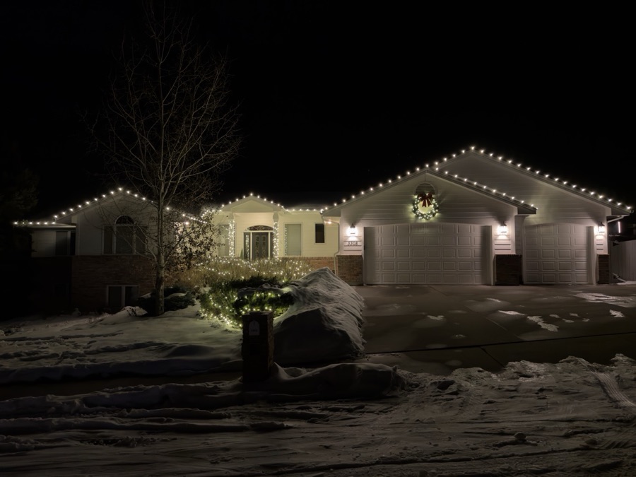 Ranch home with warm white roofline, lit shrubs and a wreath above the garage in snow