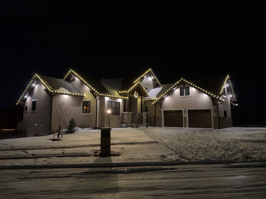 Snow-covered driveway leading to a home with warm white roofline lights on a winter evening